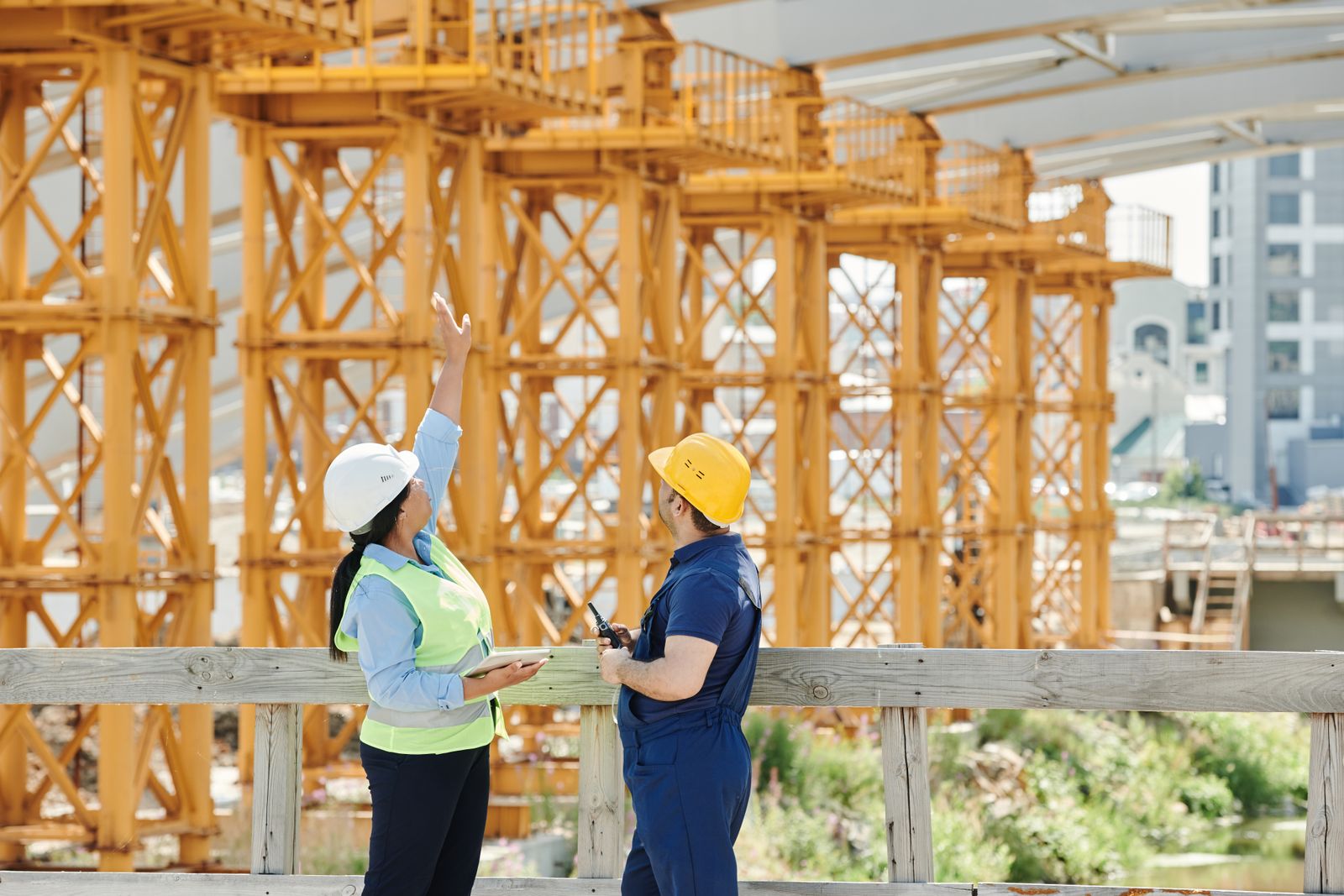 Two people in hard hats at a construction site, one holding a clipboard and the other a tablet.
