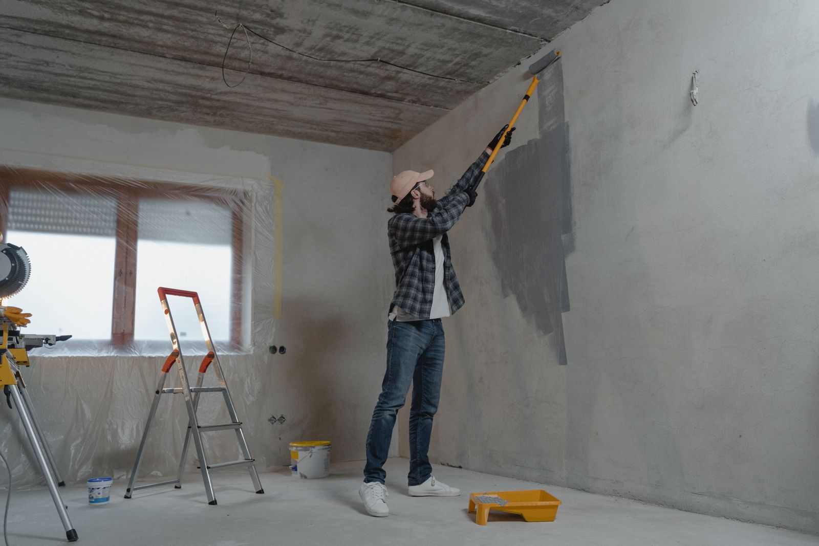 A person painting a wall in an unfinished room, with a ladder and tools nearby.